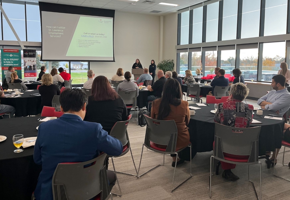 Individuals are seated at round tables listening to a presentation. At the front of the room, two individuals speak to a powerpoint presentation on a large projector screen.